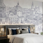 Na lewym pierwszym planie tapety widać wyraźnie Bramę Krakowską z jej wieżą zegarową, a centralnie dużą bramę miejską i liczne skrupulatnie odwzorowane kamienice Starego Miasta. Przed tapetą stoi niebieska aksamitna kanapa w kształcie litery L, geometryczny stolik kawowy i nowoczesne lampy. Całość wnętrza uzupełniają betonowe ściany, drewniane podłogi i wysoki sufit z ukrytym oświetleniem.