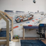 Nowoczesna aranżacja pokoju dziecięcego. Na ścianie znajduje się duża, akwarelowa tapeta dla chłopca przedstawiająca wyścigowy samochód zbudowany z klocków na torze. Wnętrze wyposażone jest w drewniane łóżko w kształcie domku, niebieski dywan, granatowe biurko z krzesłem oraz pudełko z klockami.